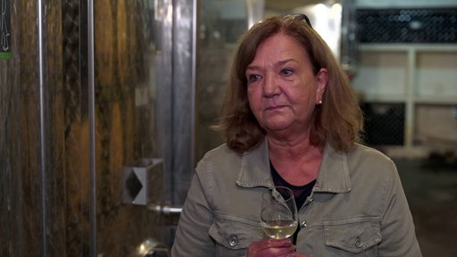 Eine mittelalte Frau mit schulterlangen Haaren steht in einem Weinkeller. In einer Hand hält sie ein Glas Weißwein.