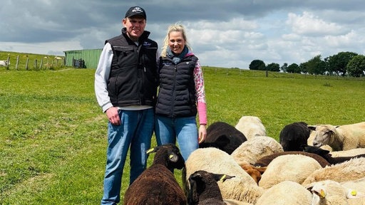 Friedel und Julia Nießen vom Lohscheidhof in Hürtgenwald stehen lächelnd auf einer grünen Wiese inmitten einer großen Schafherde. Die Schafe sind unterschiedlich gefärbt, teils schwarz, teils weiß.