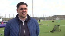 Mann mit blauer jacke steht vor einem Fußballfeld