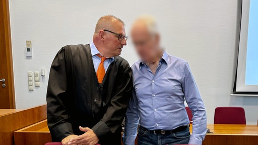Zwei Männer stehen in einem Gerichtssaal. Der Mann links trägt eine Anwaltsrobe. Der andere mann ist unkenntlich gemacht.