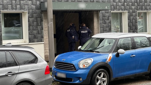 Zwei Polizisten an einer Wohnungstür, im Vordergrund ein blau-weisser Mini-Cooper