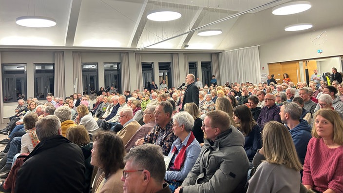 Viele Menschen sitzen in einem Saal, ein Mann steht und spricht in Mikrophon 