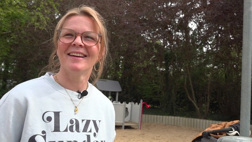 Eine lächelnde Mutter mit Brille und blondem Haar steht auf einem Spielplatz in Monheim am Rhein, im Hintergrund Bäume und Spielgeräte.
