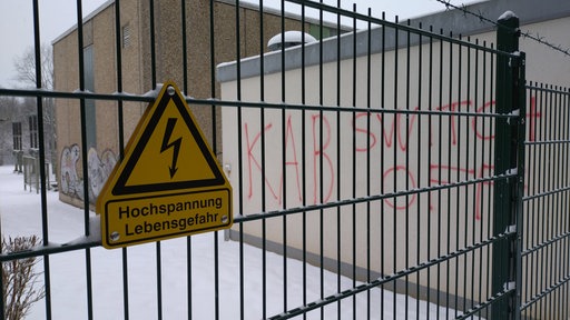 Gelbes Schild mit Blitz und Aufschrift "Hochspannung - Lebensgefahr, auf einem Metallzaun, im Hintergrund ein Trafohaus mit Graffitti