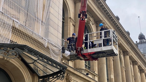 Bauarbeiter arbeiten in einem Baukran an der Fassade der Stadthalle. Die Fassade ist zum Teil mit einem Netz umhüllt.