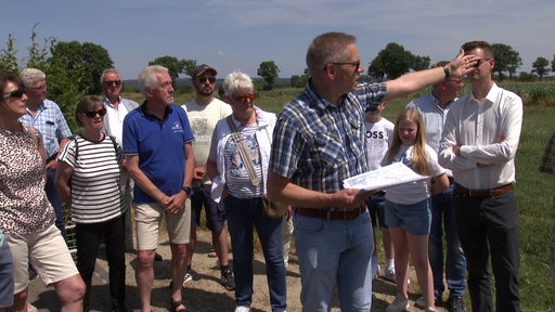 Gruppe Menschen bei einem Feld, Mann vorne hält Zettel in der Hand und zeigt in die Ferne