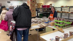 Lebensmittel stapeln sich auf einem Tisch. Menschen stehen für die Lebensmittel Schlange. Eine Frau von der Tafel schaut in die Kamera
