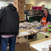 Lebensmittel stapeln sich auf einem Tisch. Menschen stehen für die Lebensmittel Schlange. Eine Frau von der Tafel schaut in die Kamera