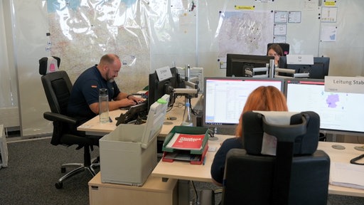 Menschen sitzen in einem Großraumbüro vor Bildschirmen. Im Hintergrund hängen Karten an der Wand. 