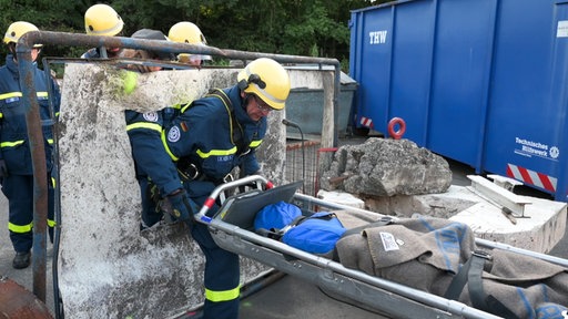 THW-Helfer in blauen Jacken tragen eine Puppe auf einer Trage durch ein Loch in einer Blechwand.