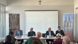Vier Männer sitzen am Tisch mit Bildern des Kölner Doms im Hintergrund.