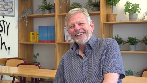 Ulrich Krapp, ehemaliger Lehrer am Otto-Hahn-Gymnasium in Monheim am Rhein, lächelt in einem Klassenzimmer mit Bücherregalen und Pflanzen