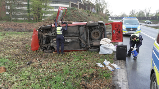 Ein rotes Auto liegt auf der Seite, um dieses herum liegen verschiedene Gegenstände. Zwei Polizisten untersuchen das Auto. 