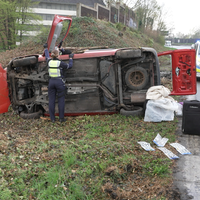 Ein rotes Auto liegt auf der Seite, um dieses herum liegen verschiedene Gegenstände. Zwei Polizisten untersuchen das Auto. 