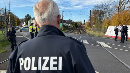 Ein Polizist steht mit dem Rücken zur Kamera. Im Hintergrund ist ein Bahnübergang dort sind weitere Polizisten und ein Einsatzfahrzeug.