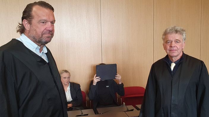 Vier Menschen in einem Gerichtssaal, einer verdeckt sein Gesicht mit einer Akte