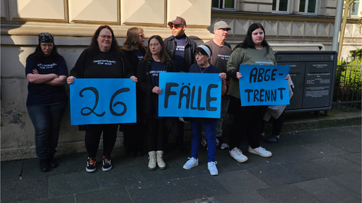 Eine kleine Gruppe von Menschen steht mit beschriebenen Plakaten vor einem Gebäude