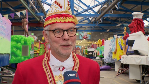 Ein Mann mit runder Brille im rot-weißen Karnevalsornat mit Kappe neben Karnevalswagen in einer Halle