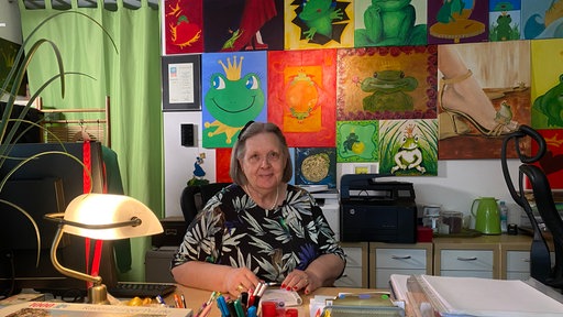 Gabriele Van den Burg sitzt in einem Büro. Im  Hintergrund Bilder von Fröschen