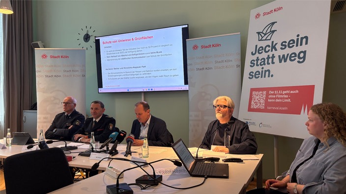 Fünf Personen sitzen vor Mikrofonen an Tischen, im Hintergrund läuft eine Präsentation