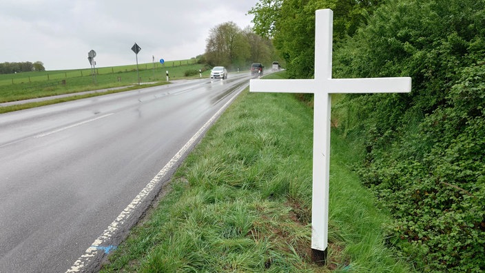 Ein weißes Holzkreuz an einer Landstraße, in der Ferne sind Autos zu sehen