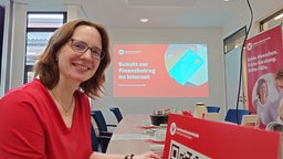 Eine Frau lächelt in die Kamera, sie sitzt an einem Schreibtisch vor einem Computer.