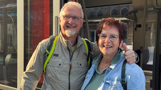 Ein Mann und eine Frau stehen Arm in Arm in der offenen Tür eines Regionalzugs und lächeln in die Kamera.