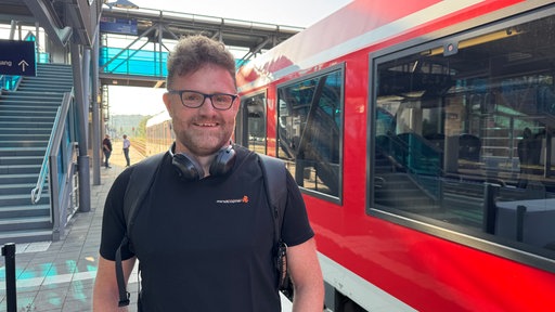 Ein Mann steht neben einem roten Zug. Er lächelt in die Kamera. Er trägt helle, kurze Haare und ein dunkles Shirt.