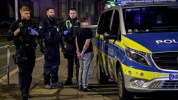 Drei Polizisten stehen mit einem Mann in Handschellen neben einem Polizeiauto