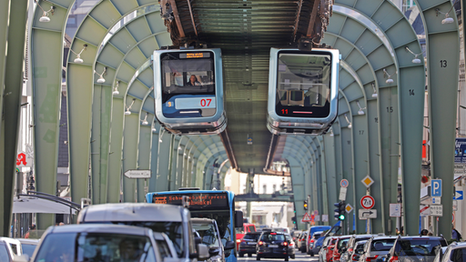 Zwei Schwebebahnen fahren auf gleicher höhe aneinander vorbei. Darunter fließt der Verkehr einer mehrspurigen Straße.