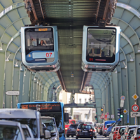 Zwei Schwebebahnen fahren auf gleicher höhe aneinander vorbei. Darunter fließt der Verkehr einer mehrspurigen Straße.
