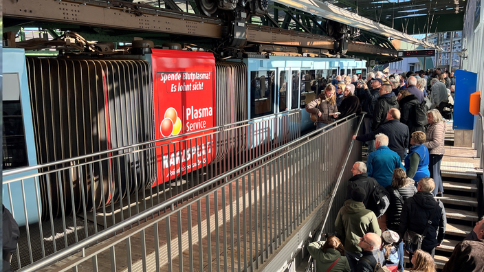 Menschen stehen auf einer Treppe in der Warteschlange, um in die Schwebebahn zu kommen. Die Türen sind noch verschlossen.