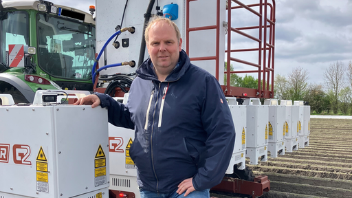 Landwirt Carsten Abenhardt steht neben seinem Traktor auf einem Feld. Sein Arm liegt auf einem der Roboter-Module am Heck des Fahrzeugs. 