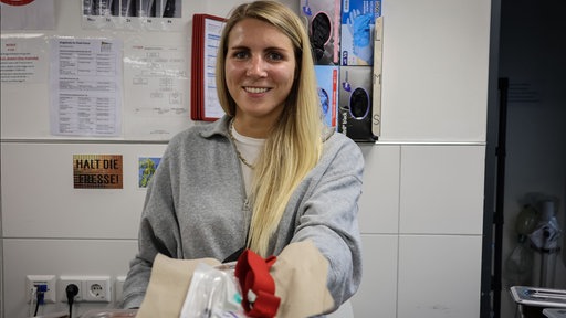 Das Bild zeigt eine Frau (Aline Dickel), die Utemsilien für eine Heroinspritze zeigt.