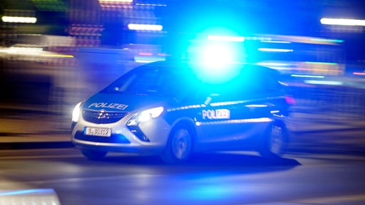 Das Bild zeigt einen Streifenwagen der Polizei mit angeschaltetem Blaulicht, der auf einer Straße fährt.