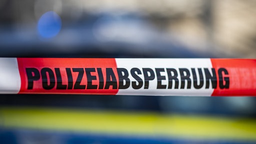 Das Foto zeigt ein Absperrband mit Polizeiwagen dahinter.