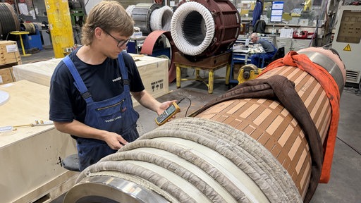Jakob Schmittel steht vor einer großen Turbine in einer Produktionshalle und schaut auf ein Messgerät.