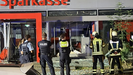 Einsatzkräfte von Polizei und Feuerwehr stehen vor einer Sparkassen-Filiale. Die Fassade ist zerstört.