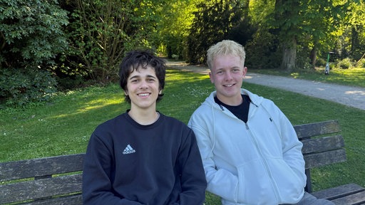 Zwei junge Männer, Kian Feldhaus und Aron Ziehn, sitzen auf einer Parkbank und lächeln in die Kamera.