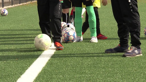 Nahaufnahme der Beine und Füße mehrerer Kinder in Fußballkleidung, die hintereinander stehen und jeweils einen Ball am Fuß haben. Daneben steht ein Mann in Jogginghose. 