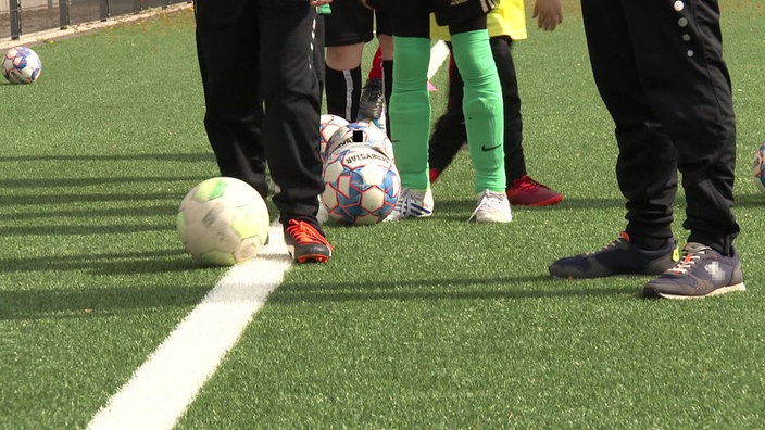 Nahaufnahme der Beine und Füße mehrerer Kinder in Fußballkleidung, die hintereinander stehen und jeweils einen Ball am Fuß haben. Daneben steht ein Mann in Jogginghose. 