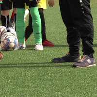 Nahaufnahme der Beine und Füße mehrerer Kinder in Fußballkleidung, die hintereinander stehen und jeweils einen Ball am Fuß haben. Daneben steht ein Mann in Jogginghose. 