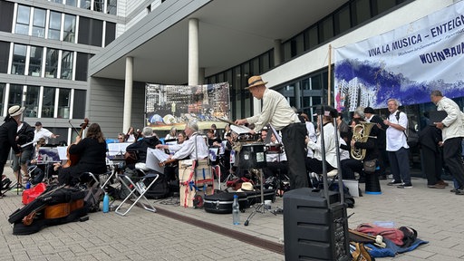 Mehere Musikerinnen und Musiker sitzen mit ihren Instrumenten vor dem Eingang der Vonovia-Zentrale in Bochum, vor dem Fenster hängt ein weiß-blaues Plakat mit der Aufschrift "Viva la Musica"