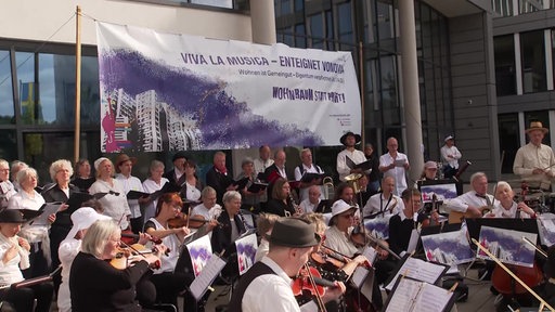 Mehrere Musikerinnen und Musiker sind vor der Vonovia-Zentrale und spielen mit ihren Instrumenten, im Hintergrund ist ein Banner zu sehen mit der Aufschrift: "Viva la Musica - enteignet Vonovia"