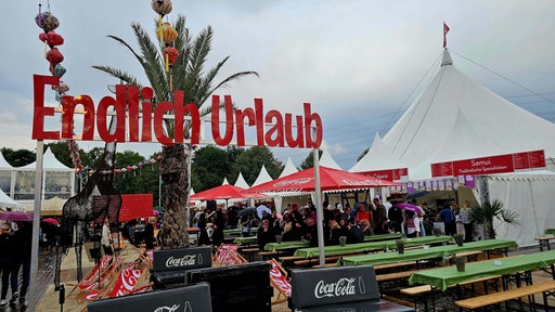 Über Bierbänken und zwischen Festivalzelten steht ein Banner mit der Aufschrift "Endlich Urlaub" 