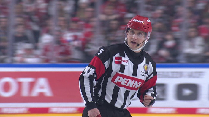 Andre Schrader läuft im Schiri-Trikot über das Eis und spricht in ein Mikrofon.