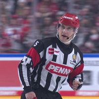 Andre Schrader läuft im Schiri-Trikot über das Eis und spricht in ein Mikrofon.