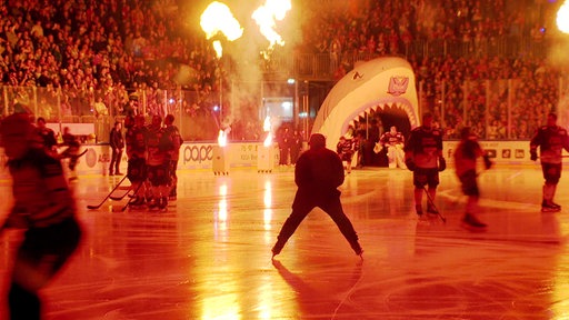 Mehrere Eishockeyspieler laufen über das Eis. Im Hintergrund ist die Tribüne mit Zuschauern zu sehen. Es startet eine Feuershow. 