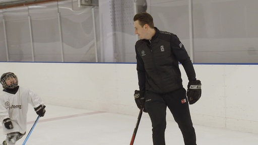 Andre Schrader läuft mit Hockeyschläger in der Hand über das Eis. An der Seite steht ein Kind in Eishockeykleidung mit Schläger in der Hand. 