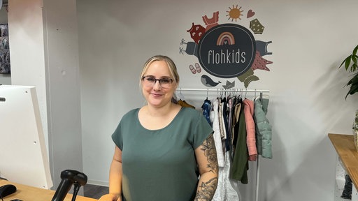 Sarah Degen steht lächelnd hinter der Kasse, ein "flohkids" Wand-Tattoo und eine Kleiderstange hinter ihr.